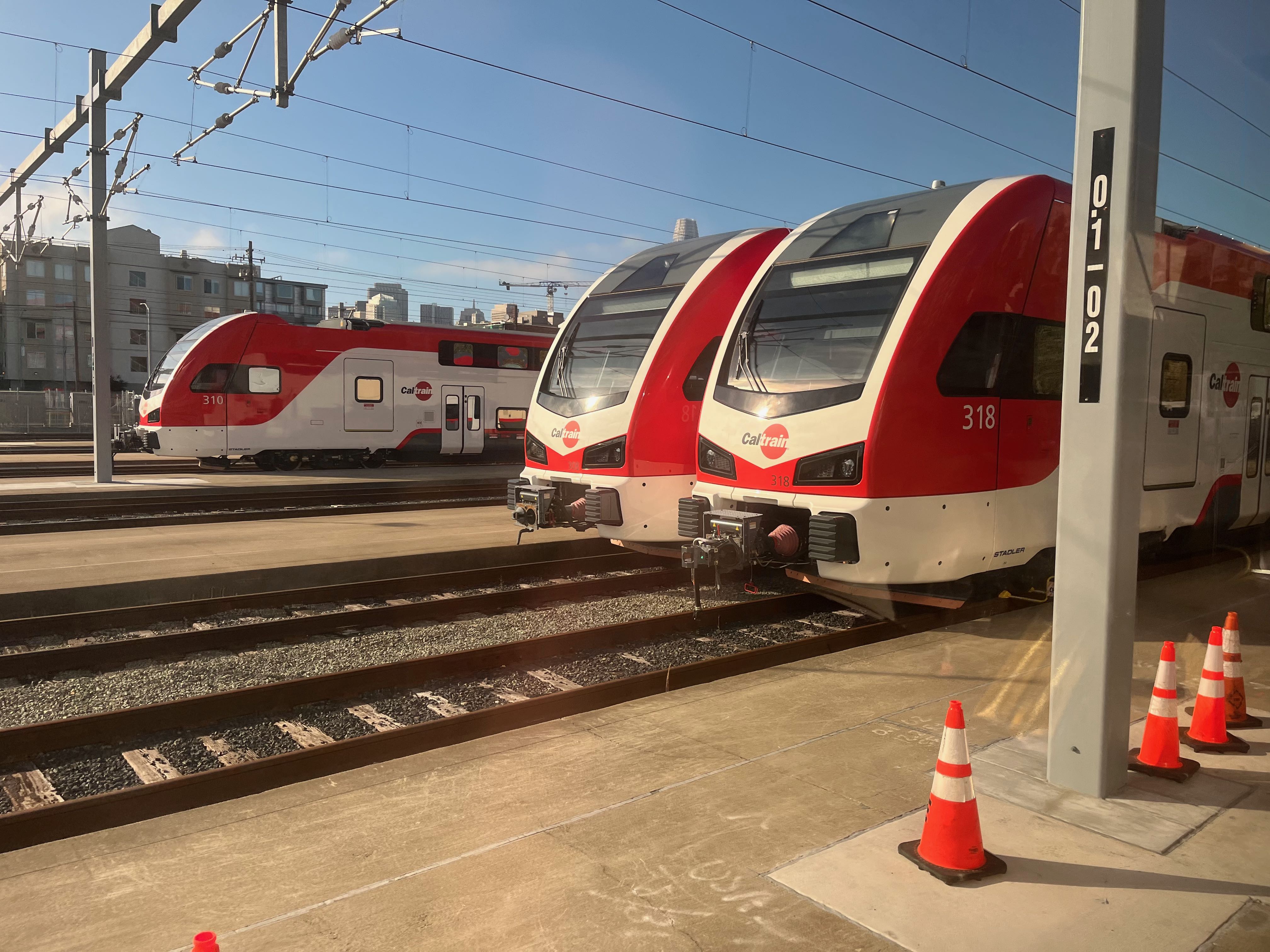 A view looking across the tracks at the San Francisco Caltrain station, showing the ends of three Stadler electric multiple unit trains with sunlight shining on them. A collection of three orange cones sits near the center of an island boarding platform in the foreground. A large pole with cantilevered structures holds up overhead wires above the tracks.