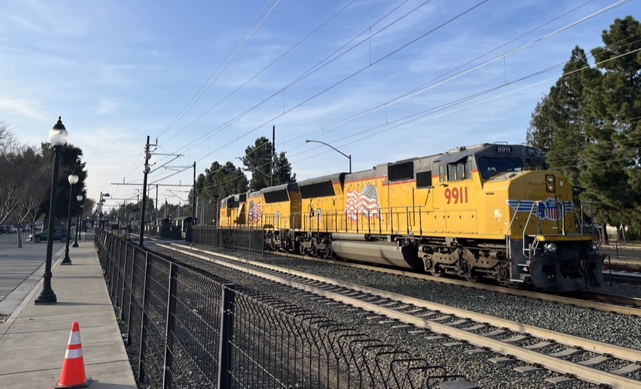 A view looking north along the Caltrain railroad from beyond a fence on the west side of the tracks. A Union Pacific freight locomotive is on the northbound track. A construction cone is on the ground at the bottom left corner of the photo.